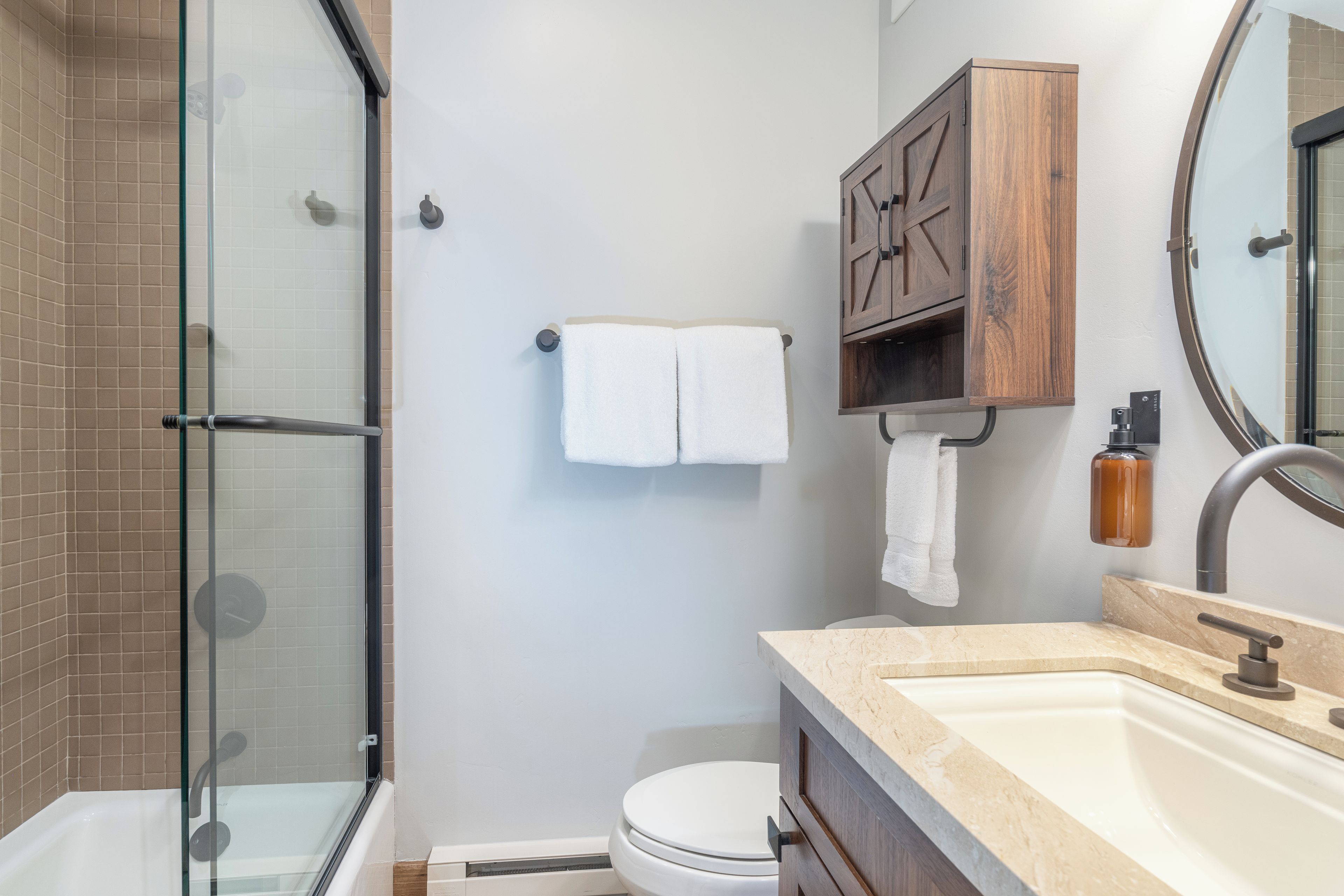 A bathroom with a glass shower enclosure, a toilet, a sink with a marble countertop, and a wooden medicine cabinet.