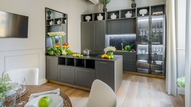 Een moderne keuken met grijze kastjes, een centraal kookeiland met vers fruit en groenten, en een eetgedeelte met een tafel gedekt voor twee personen.