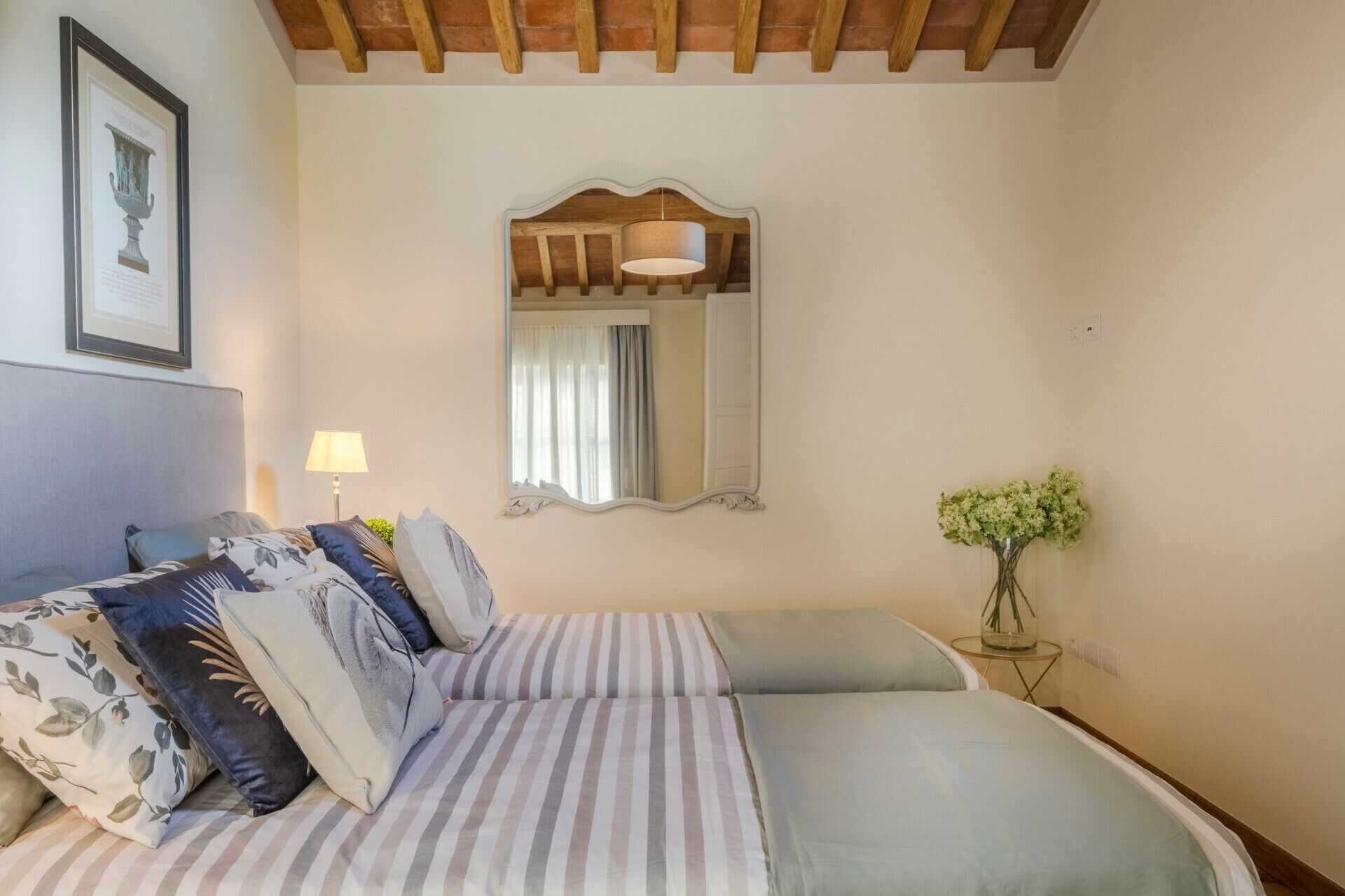 Une chambre à coucher avec un lit, une lampe de chevet, un miroir et un vase rempli de fleurs.