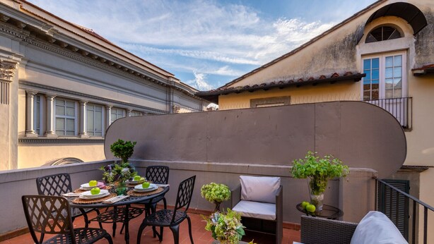 An outdoor patio with a round table set for dining, surrounded by wicker chairs. There are potted plants and a small table with fruits and vegetables. - Elegant Modern Apartment with Terrace in Historic Lucca (Lucca)