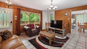 A cozy living room with a leather sofa, a wooden coffee table, a TV, and a large window with a view of greenery. - Havre de Détente & Spa (Sainte-Lucie-des-Laurentides)