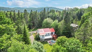 A house surrounded by dense forest with a red roof, a visible driveway, and a small patio area with outdoor furniture. - Havre de Détente & Spa (Sainte-Lucie-des-Laurentides)