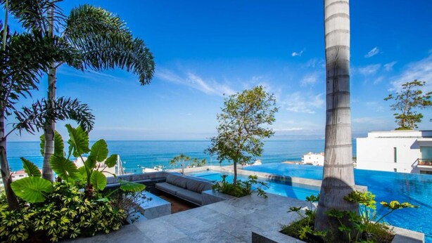 Un área de piscina moderna con vista al océano, rodeada de plantas tropicales y una palmera alta.
