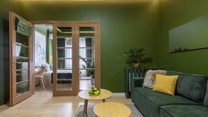 Ein Zimmer mit grüner Wand, einem grünen Sofa mit gelbem Kissen, einem Holztisch und einer Tür zu einem anderen Raum.