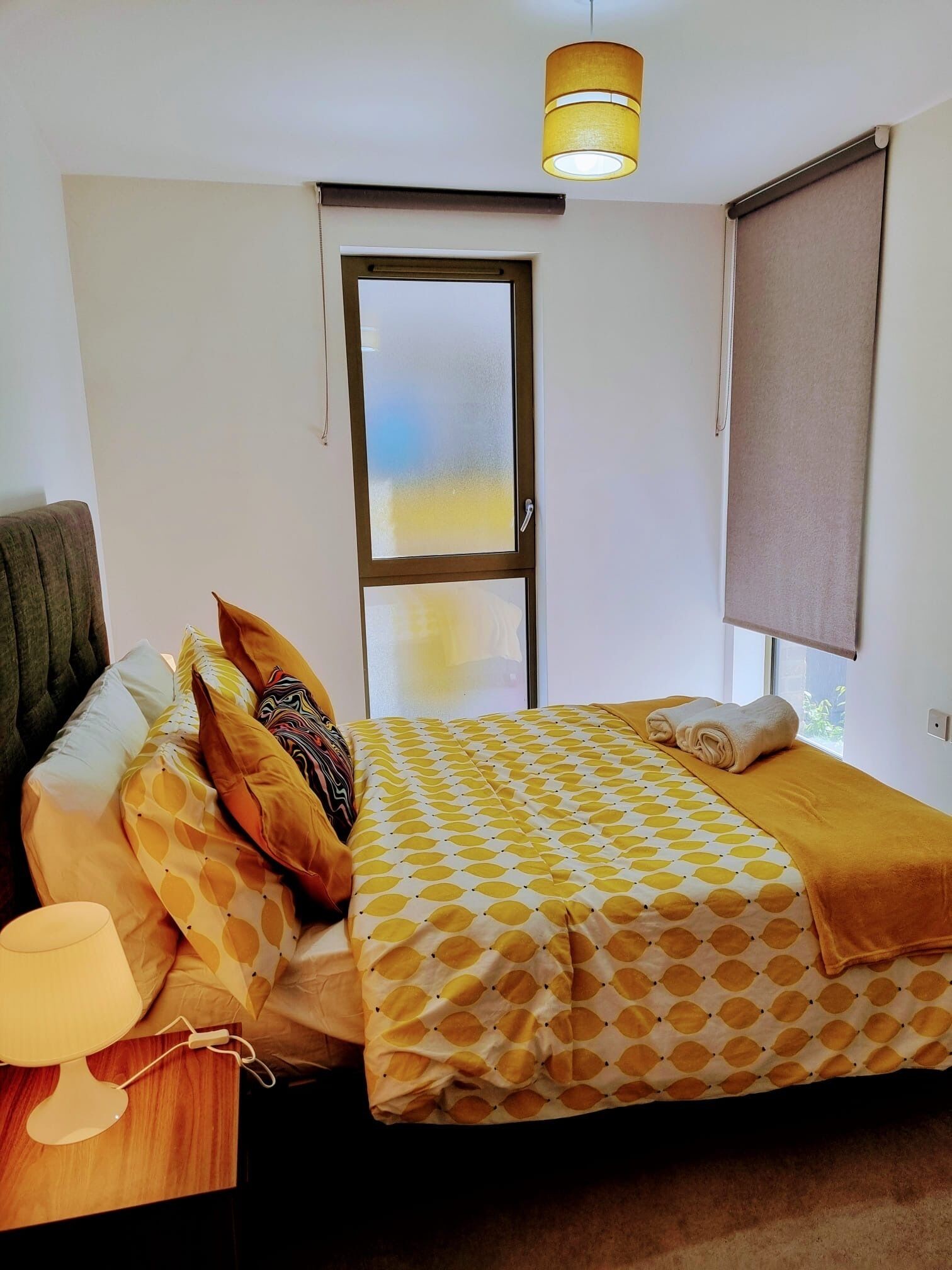 A bedroom with a bed, pillows, a bedside table with a lamp, a window with a blind, and a door with frosted glass.