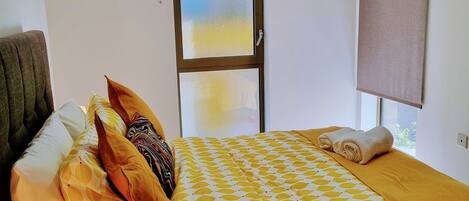 A bedroom with a bed, pillows, a bedside table with a lamp, a window with a blind, and a door with frosted glass.