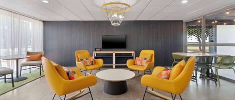 A modern meeting room with yellow rocking chairs, a central table, and a large flat-screen TV on the wall.