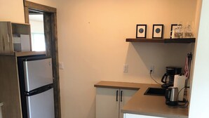 A compact kitchen with a refrigerator, a coffee maker, and a shelf with framed pictures.