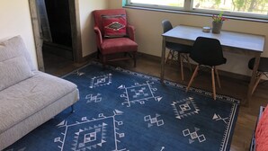 A room with a sofa, a red armchair with a patterned cushion, a small table with two chairs, and a window with a view of trees and construction outside.