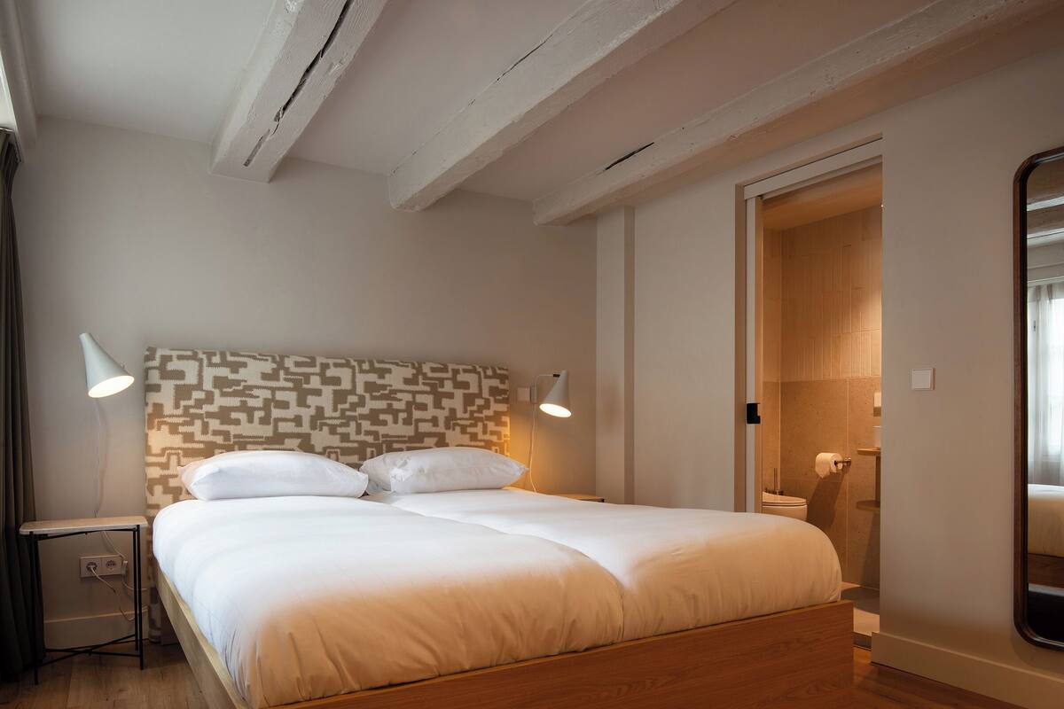 Une chambre à coucher avec une tête de lit à motifs, un cadre de lit en bois, deux lampes de chevet et une porte donnant sur une salle de bain.