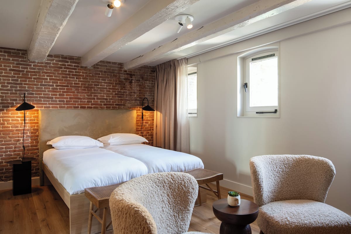 Une chambre d’hôtel avec un mur en briques, deux lits, une petite table, deux chaises et une fenêtre.