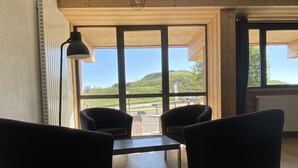 Una habitación con dos sillones negros, una mesita, una lámpara de pie y ventanas amplias que ofrecen vistas a un paisaje verde.
