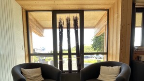 Una habitación moderna con dos sillones negros, una mesa de centro con troncos de madera y un ventanal con vistas a un aparcamiento y zonas verdes.