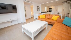 Ein modernes Wohnzimmer mit einem weißen Couchtisch, einem orangefarbenen Sofa mit gelben Kissen und einem an der Wand montierten Flachbildfernseher.