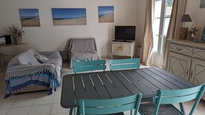 Um quarto aconchegante com uma mesa de jantar, cadeiras azul-turquesa, um sofá com manta listrada, uma TV sobre um móvel branco e arte decorativa com tema praiano.