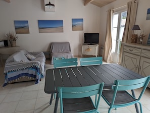 Et hyggeligt rum med et spisebord, turkise stole, en sofa med et stribet tæppe, et fjernsyn på et hvidt skab og vægkunst med strandtema.