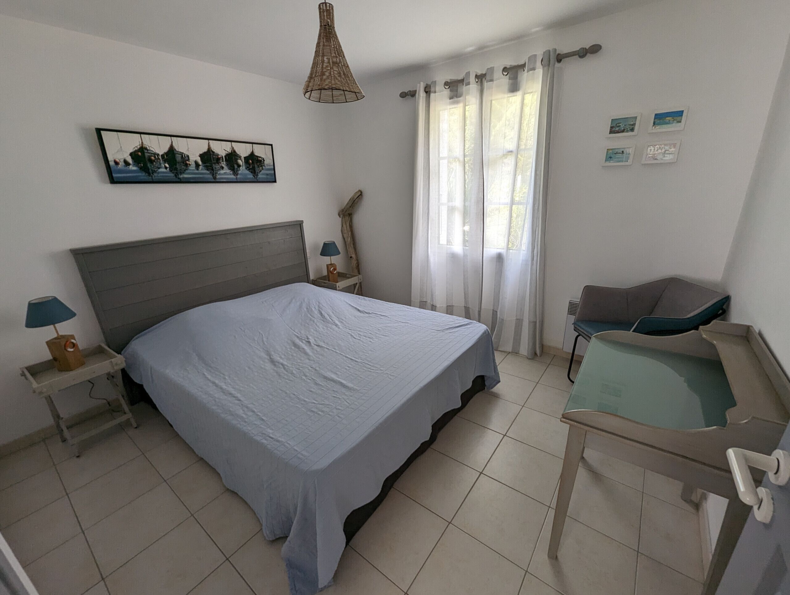 Ein Schlafzimmer mit einem Bett, einem Nachttisch, einer Lampe, einem Sessel, einem Fenster mit Vorhängen und einem Wandbild.