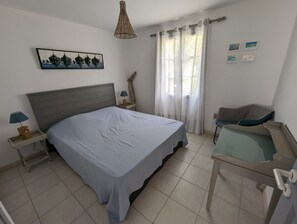 Ein Schlafzimmer mit einem Bett, einem Nachttisch, einer Lampe, einem Sessel, einem Fenster mit Vorhängen und einem Wandbild.
