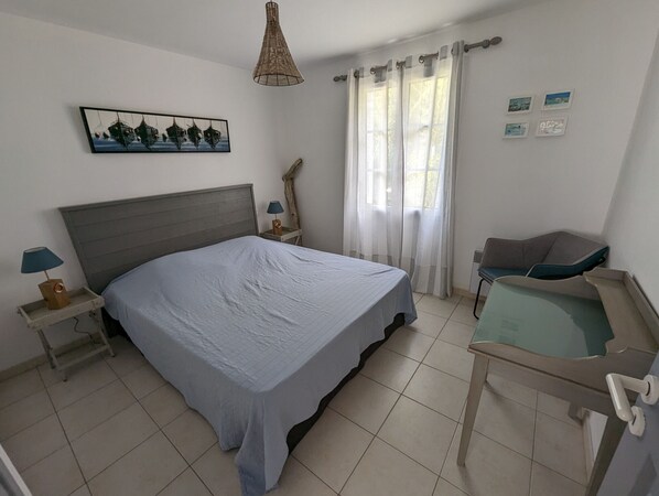 Une chambre à coucher comprenant un lit, une table de chevet, une lampe, une chaise, une fenêtre avec des rideaux et un tableau accroché au mur.