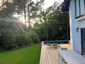 Une terrasse en bois avec des chaises longues et une piscine, entourée d’une végétation luxuriante et d’arbres.