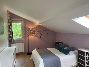 Une chambre avec un plafond en pente, un lit avec une literie blanche et des coussins turquoise, une lampe sur pied, un radiateur et une fenêtre avec des rideaux.