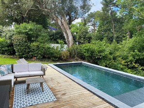 Une piscine rectangulaire dotée d’une terrasse en bois et d’un espace détente avec un canapé et un tapis à motifs.