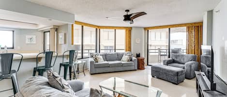 A spacious living room with a grey sofa set, a glass coffee table, and a ceiling fan.