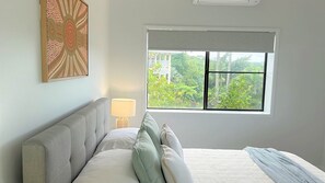 A bedroom with a bed, pillows, a nightstand, a lamp, and a window with a view of greenery.