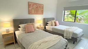 A bedroom with two beds, nightstands, a wall-mounted artwork, and a window with a view of greenery.