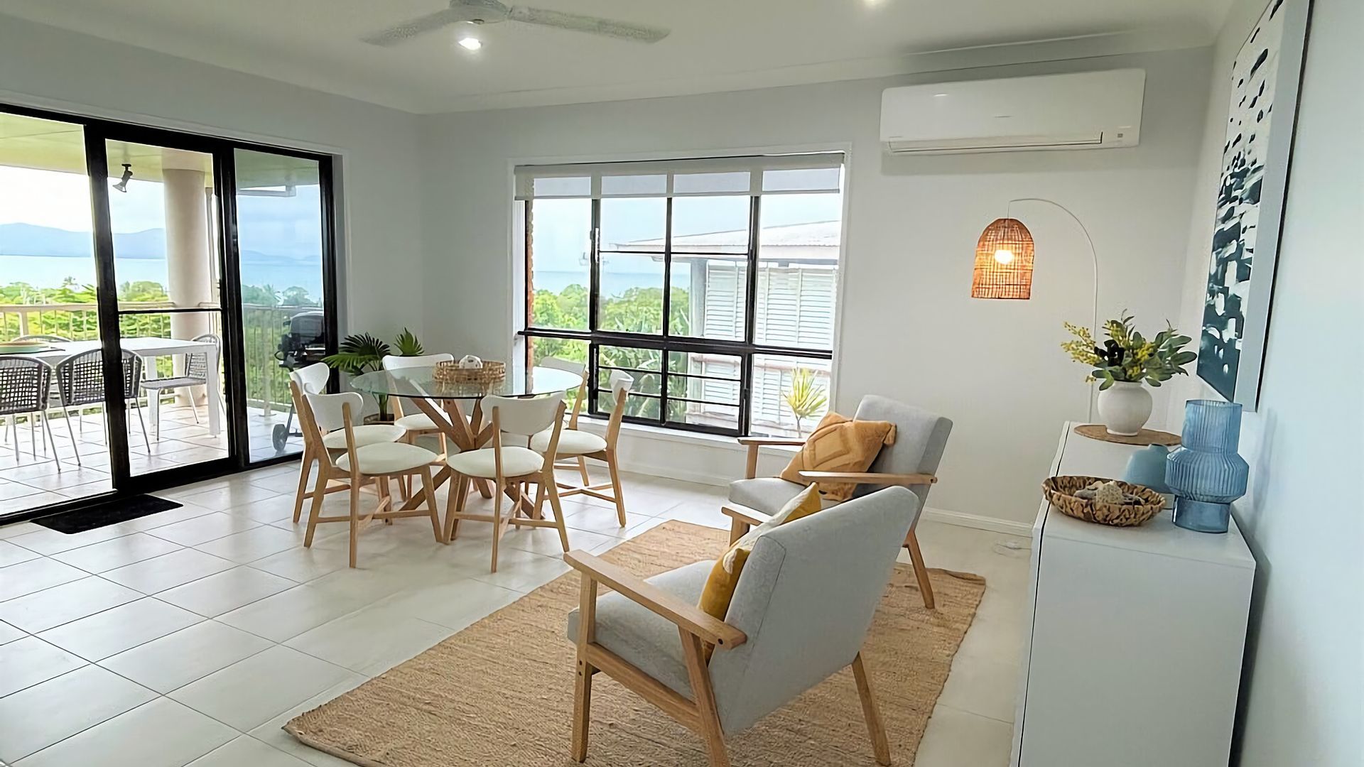 A modern living room with a glass table, white chairs, a sofa, and a view of the outdoors.