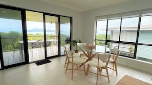 Comedor con mesa y sillas de vidrio, ventanas amplias y vista a zonas verdes y montañas.