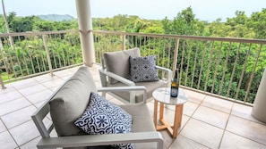 A balcony with a sofa and a small table, a bottle of champagne, and a view of lush greenery.