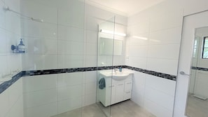 Un baño moderno con azulejos blancos, cerramiento de ducha de vidrio y un lavamanos blanco con detalle en azulejo negro.