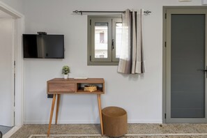 A room with a TV mounted on the wall, a small wooden desk with a basket, a round stool, and a door with a window. - Makris Gialos Lemon Cottage in Lassi Kefalonia (Kefalonia)
