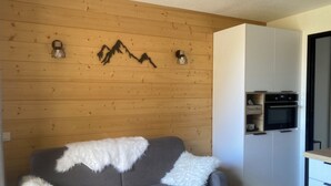 A cozy living area with a grey sofa, white fluffy throw, wooden wall paneling, and a built-in kitchen with a white cabinet and a black oven.