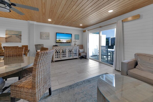 A spacious living area with a large glass table, wicker chairs, a white cabinet, and a television displaying a beach scene.