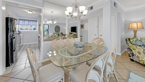Een eetruimte met een glazen tafel en witte stoelen, een kroonluchter en uitzicht op de keuken.