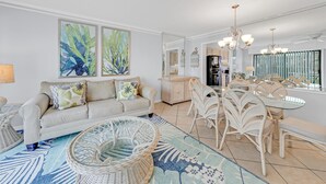Een moderne woonkamer met een bank, rieten stoelen, een eettafel met glazen blad en kunstwerken aan de wanden.