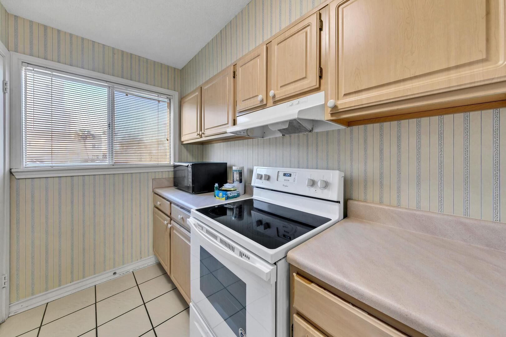 A compact kitchen with a microwave, stove, and oven. There is a window with blinds, and the walls have a striped pattern.