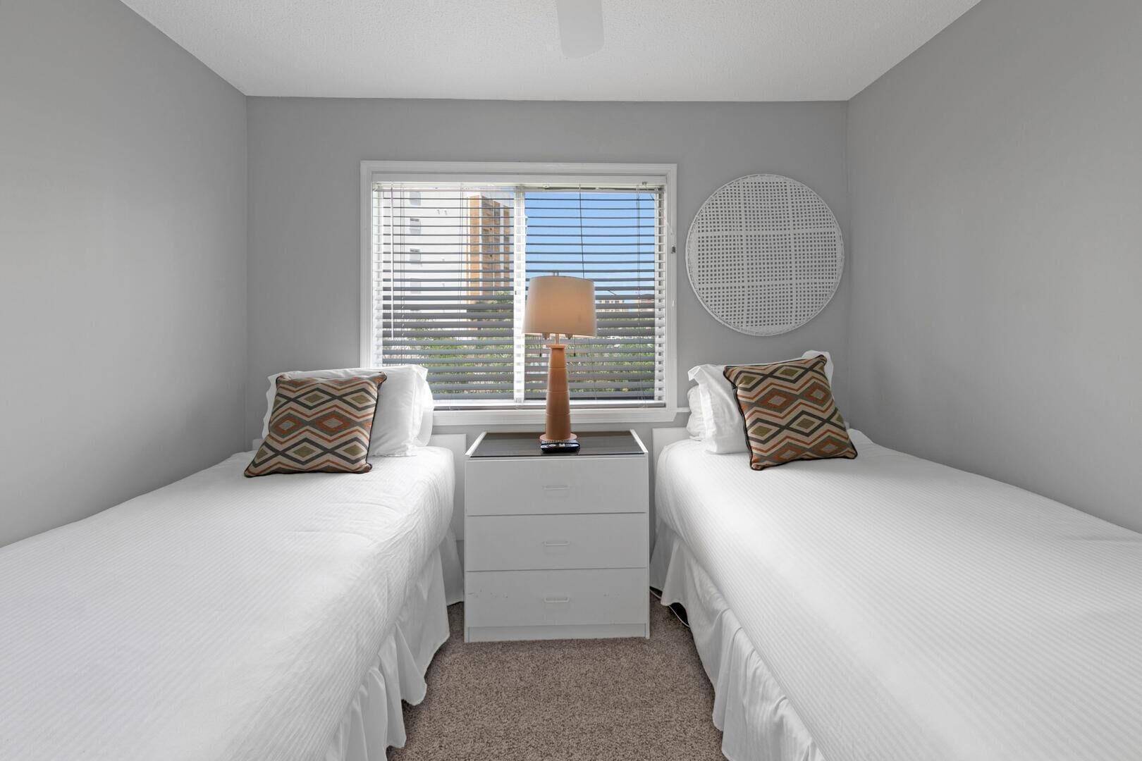 Une petite chambre propre, avec deux lits simples, une table de chevet avec une lampe et une fenêtre avec des stores.