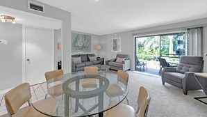 Ein modernes Wohnzimmer mit einem Glastisch, umgeben von Stühlen, einem grauen Sofagarnitur und einem Balkon mit Gartenmöbeln.