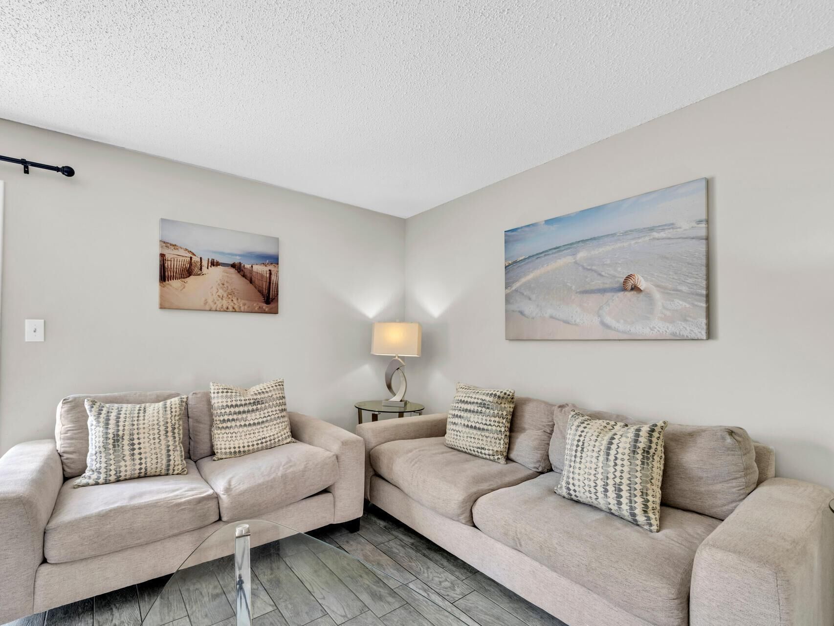 A living room with two beige sofas, patterned cushions, a side table with a lamp, and two beach-themed wall art pieces.
