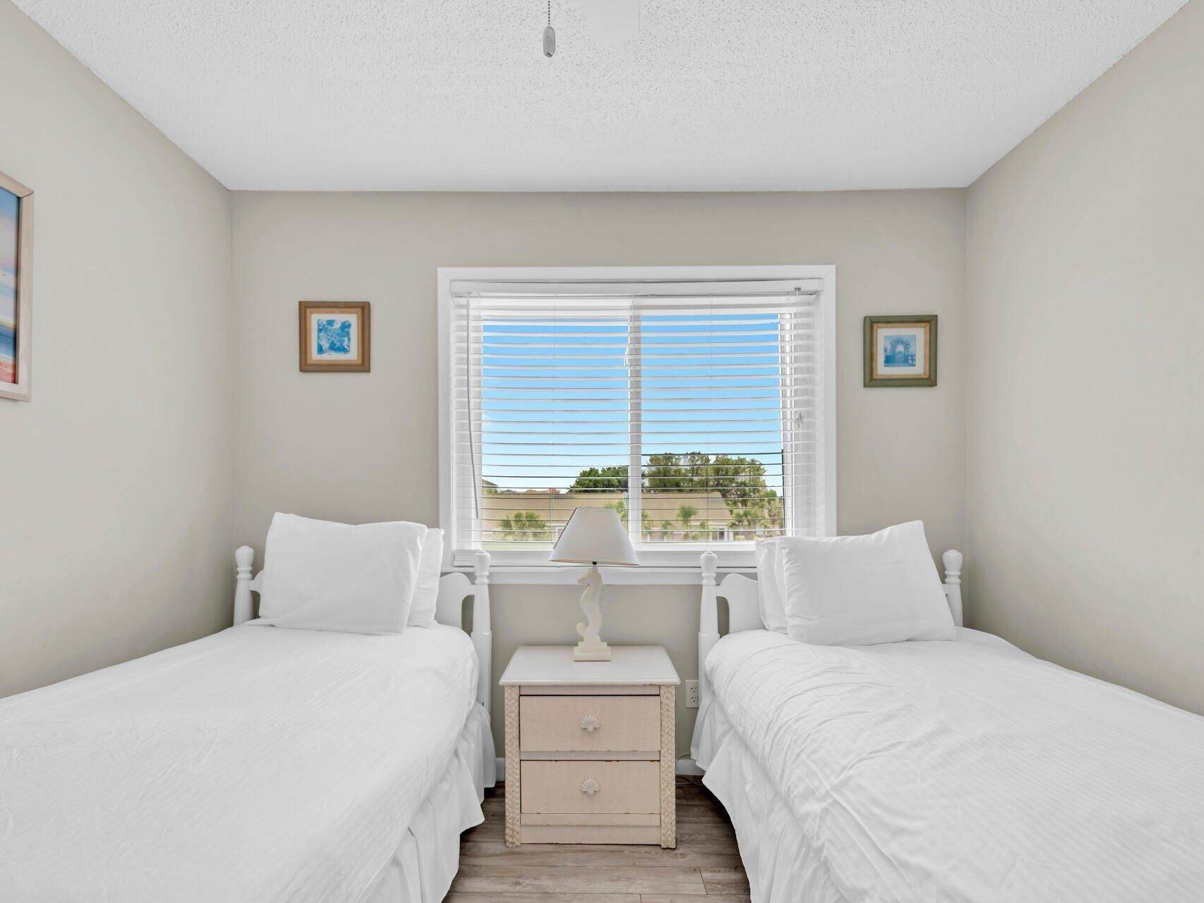 A bedroom with two beds, a window with blinds, and a nightstand with a lamp.