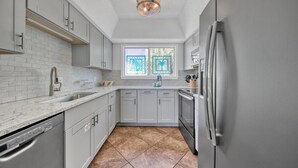 A kitchen with grey cabinets, a stainless steel refrigerator, and a window with a view of palm trees. - Southbay by the Gulf 124 (Destin)