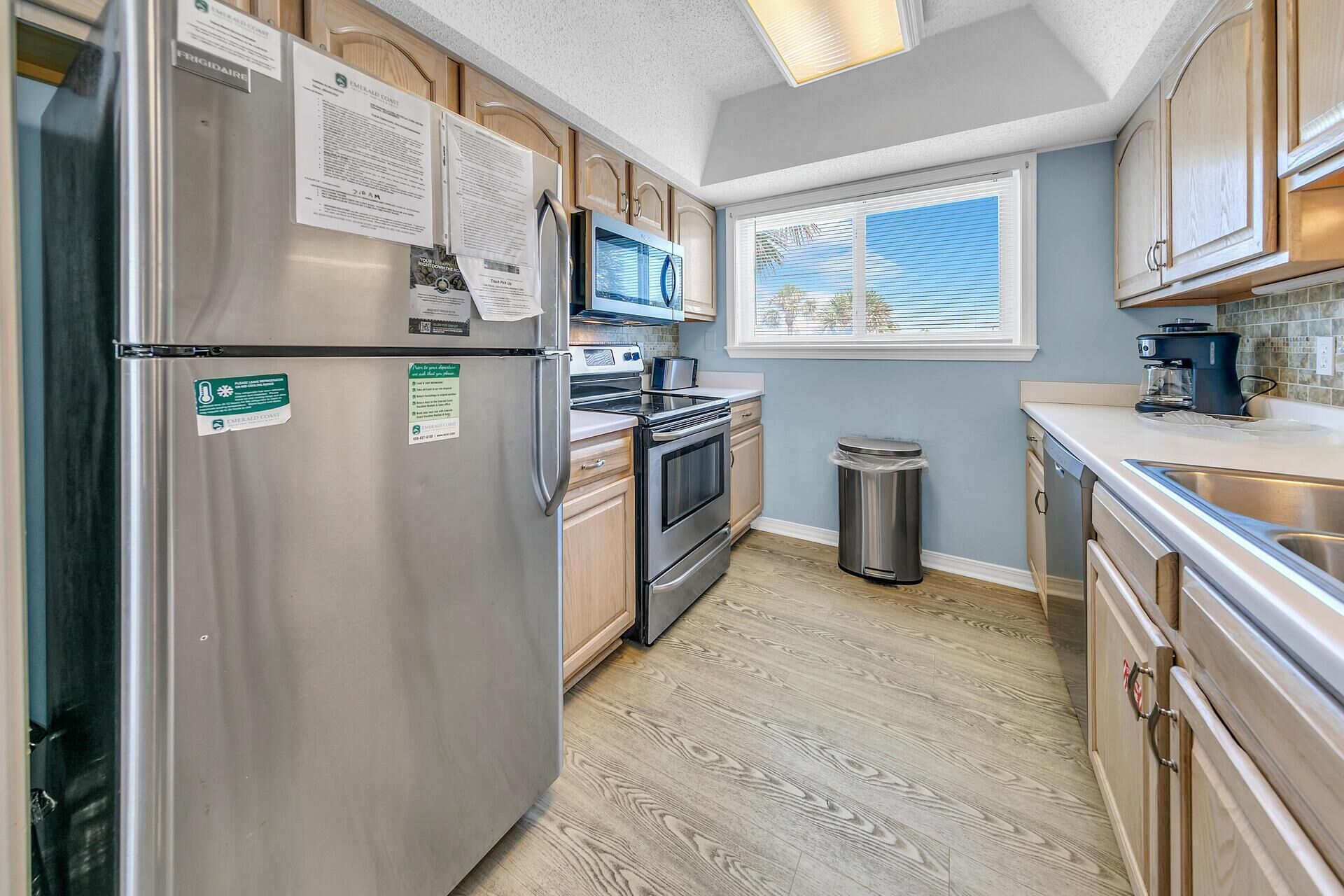 A compact kitchen with a stainless steel refrigerator, a microwave, an oven, and a sink. There are wooden cabinets and a window with blinds.
