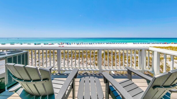 Ein Balkon mit zwei Stühlen und einem Tisch mit Blick auf einen Strand, auf dem Sonnenschirme und Personen sind.