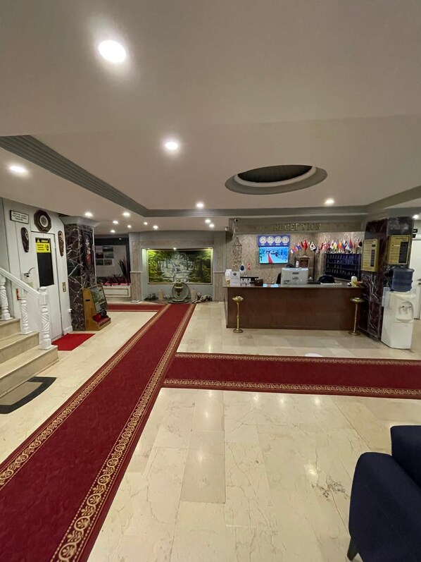Zona de recepción de un hotel con suelo de mármol, una alfombra roja que conduce a la entrada, un mostrador de recepción y una pared con banderas y un reloj.
