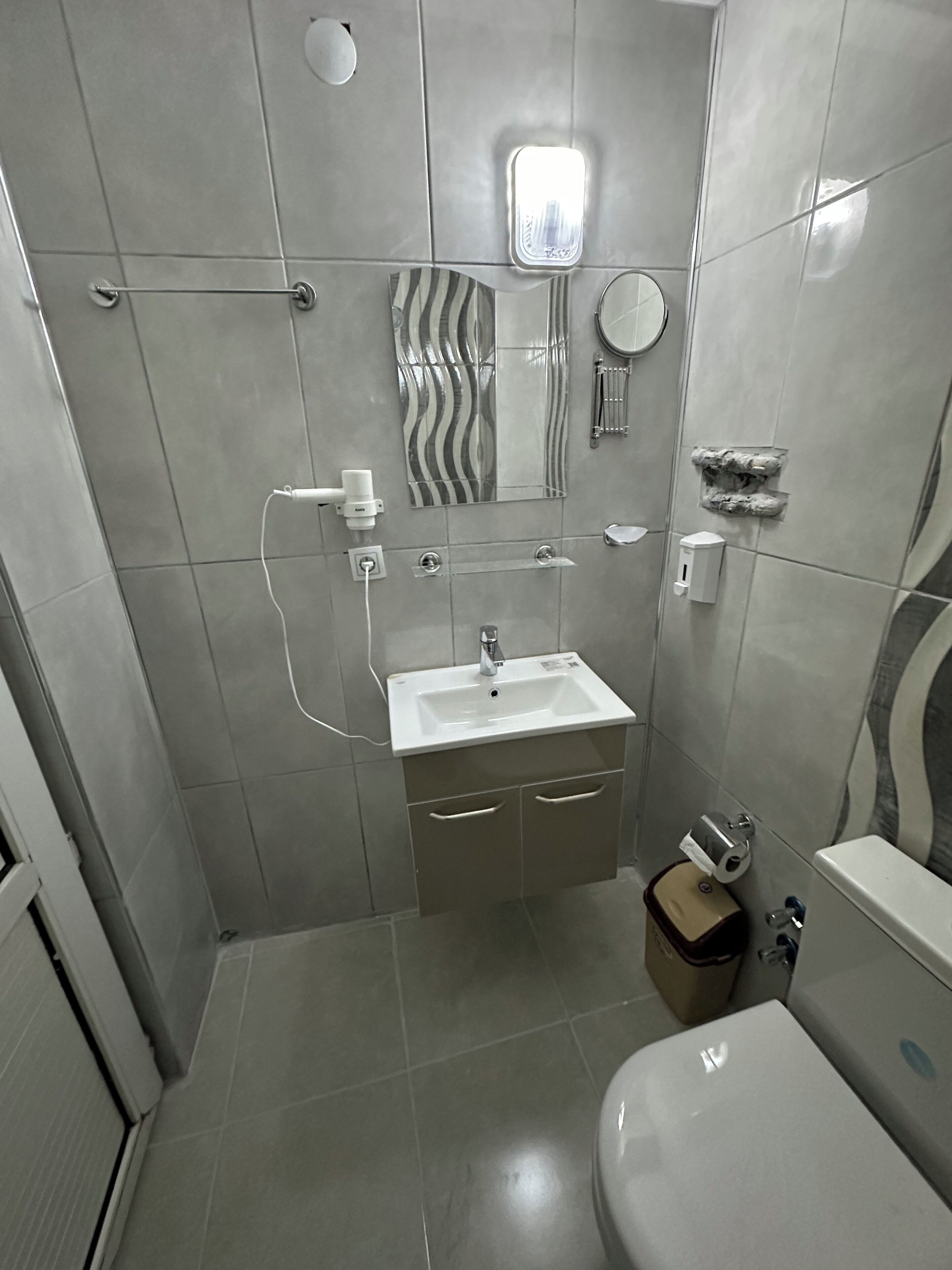 A modern bathroom with a white sink, mirror, and toilet. There is a towel rack, a soap dispenser, and a small trash bin.