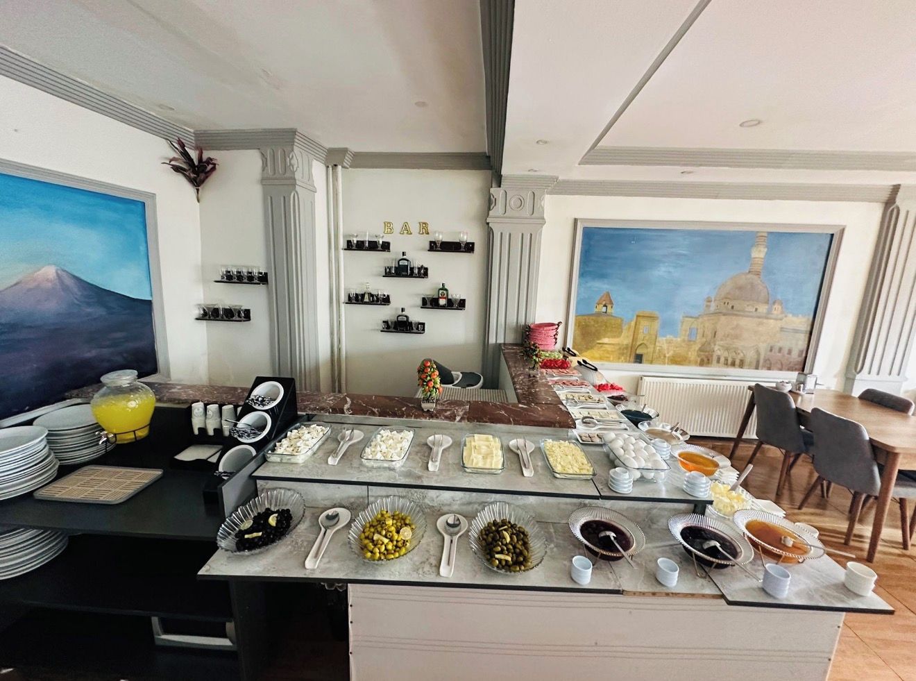 Buffet de café da manhã com diversos alimentos e utensílios, área de bar com garrafas e copos, e uma grande janela com vista para um edifício.