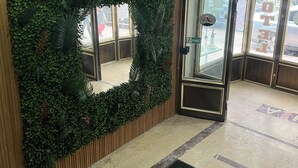 A hotel entrance with a green artificial hedge, a glass door with a sign, and a doormat with the hotel's name.
